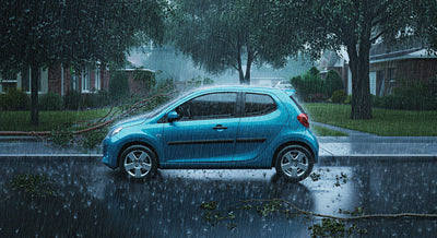 A small blue car in a suburban neighborhood during a heavy rain storm with limbs fallen around it.