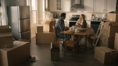 A couple sitting in their kitchen with partially unpacked boxes around them.