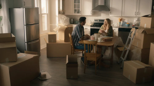 A couple sitting in their kitchen with partially unpacked boxes around them.