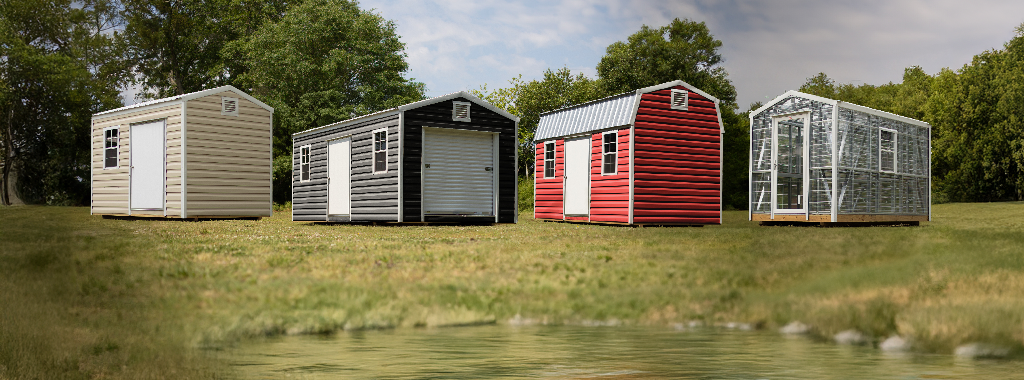 Four small storage sheds in different colors lined up by a body of water with trees in the background.
